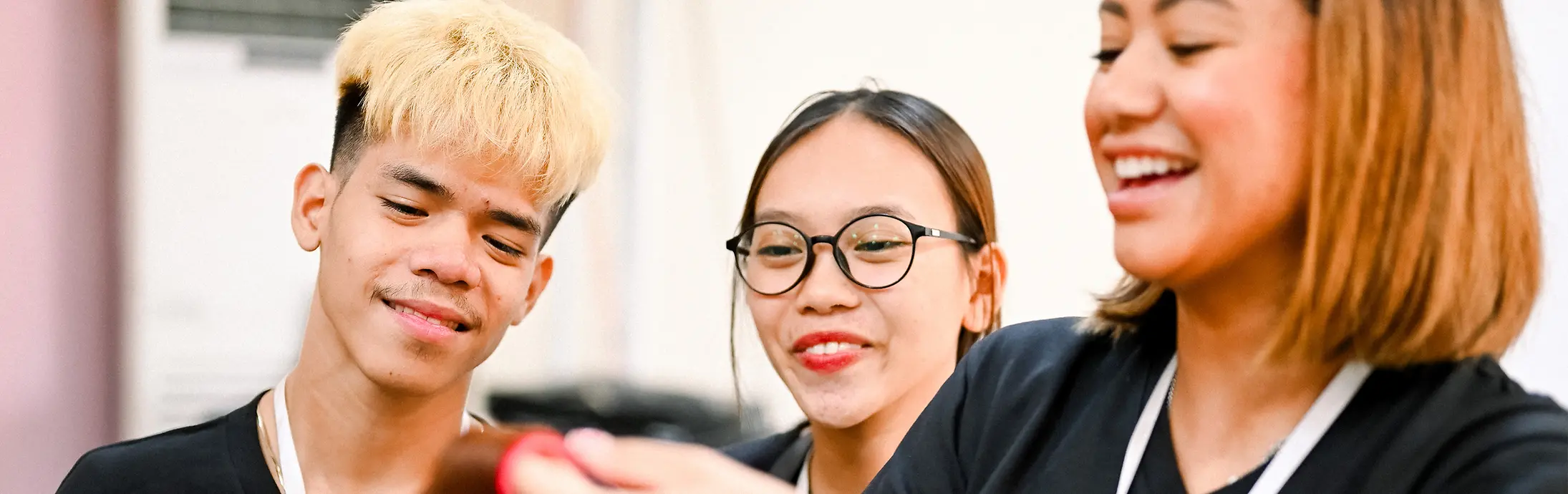 Two smiling young adults watch attentively as a Shaping Futures trainer styles the hair on a practice mannequin.