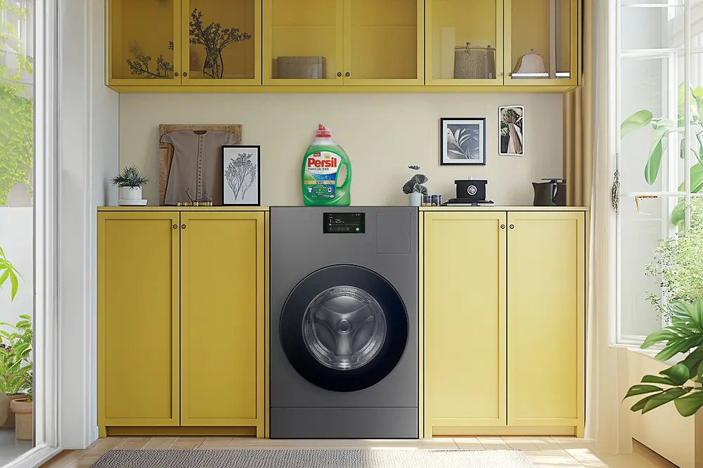 A Washing machine in the middle of a living room with Persil laundry detergent standing on top of it.