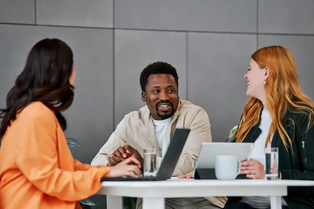 Three people – two women and one man – are sitting at a table in a modern work environment, engaged in a lively conversation. Laptops and coffee cups are on the table.