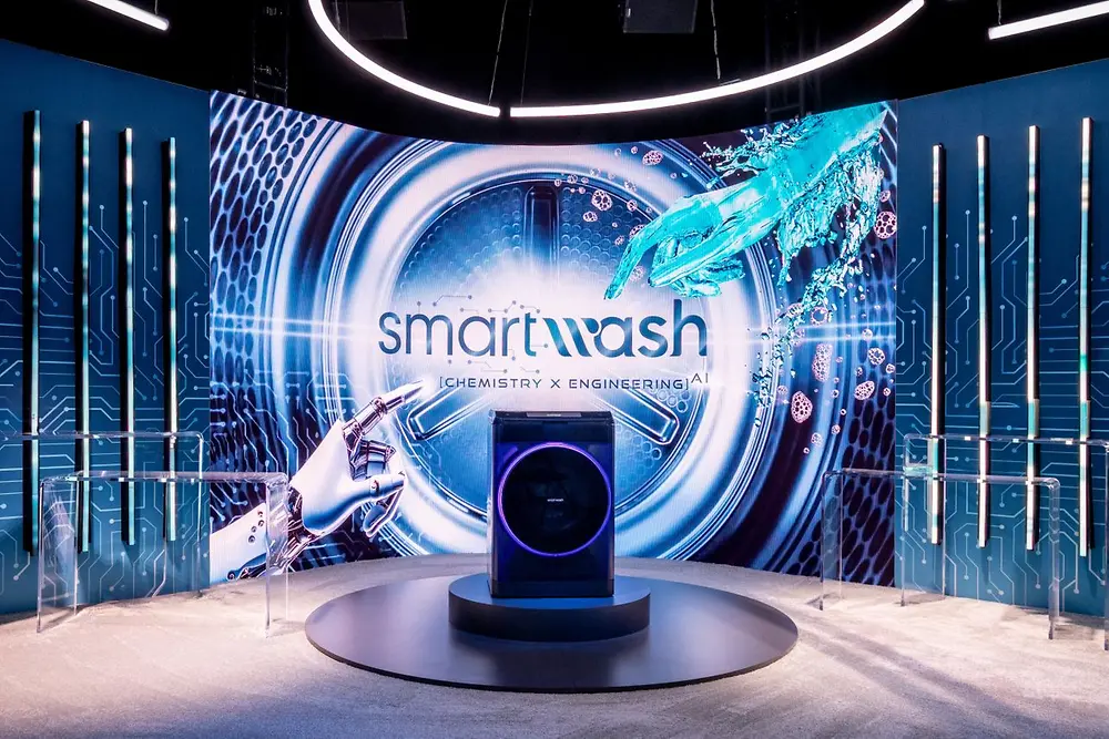 A futuristic scene features a washing machine on a small, round stage at the center. Behind it, a large screen displays a close-up of a washing machine drum and the writing 