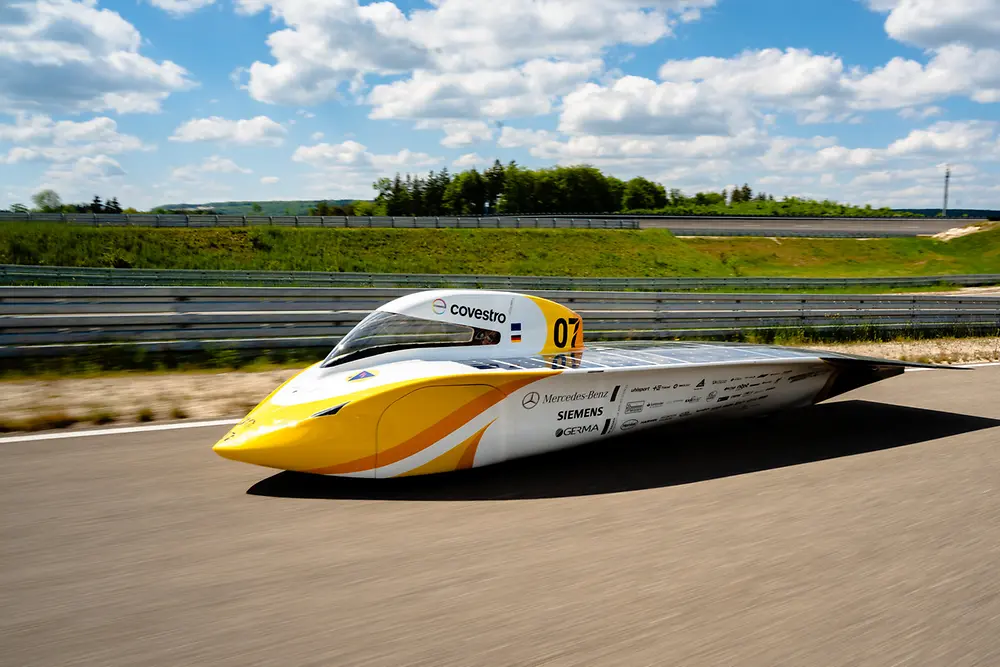 The yellow and white solar car “Sonnenwagen” drives on a racetrack, with green landscape and blue sky in the background.