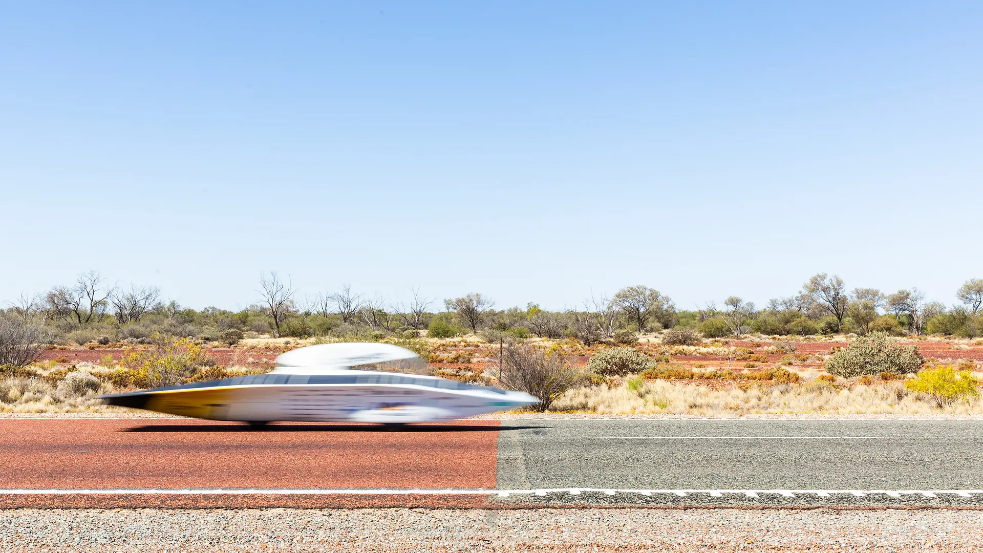 The solar car is driving at high speed on a road in a desert landscape. The vehicle appears blurred due to motion. In the background, there are dry bushes and trees under a clear blue sky.