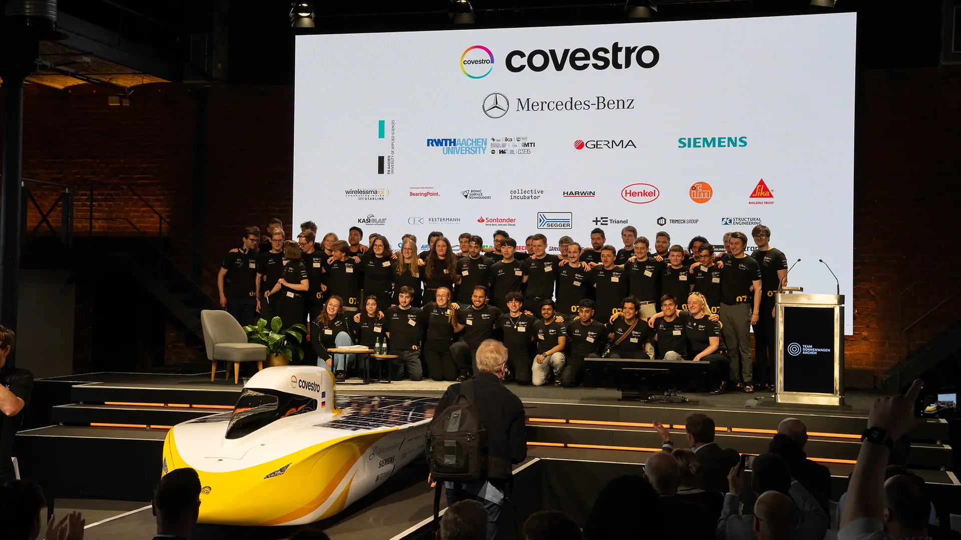 Students from RWTH Aachen University pose on a stage with the Sonnenwagen, their self-built solar car. Sponsor logos are displayed on a large screen in the background.
