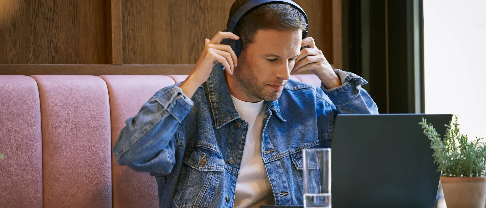 A man wearing a denim jacket and headphones is sitting at a table in a cafe, working on a laptop. There is a glass of water and a potted plant on the table.