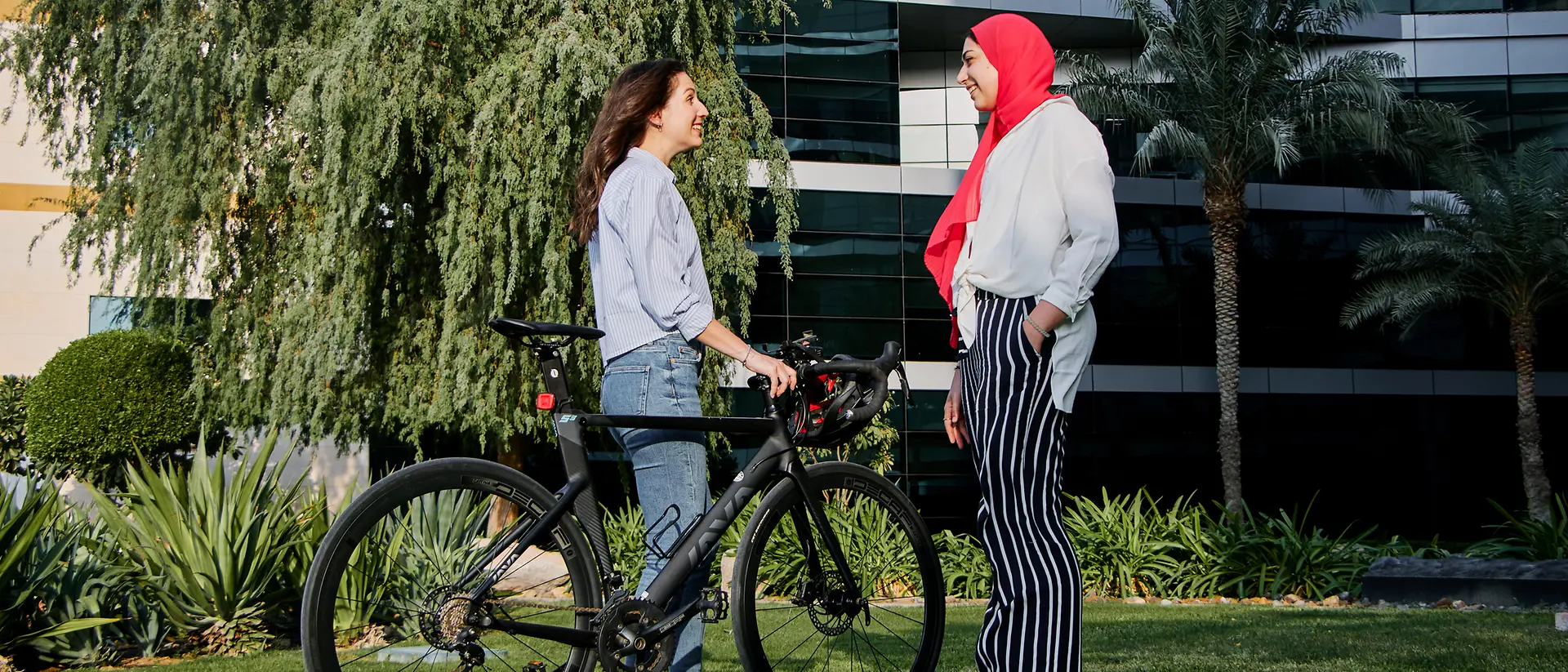 A photo of two women standing together in a green space in front of an office building with one of them holding her bike and helmet. 