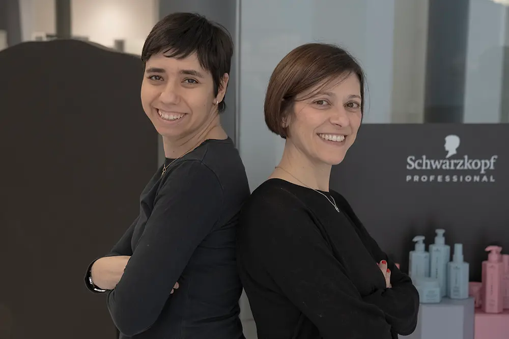Noemí and her former trainer Meritxell stand back-to-back in a salon, smiling sideways toward the camera. 