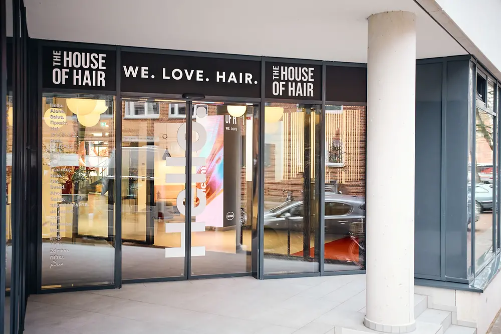 The entrance area of the House of Hair with large glass windows and doors. Above the entrance, the words “WE. LOVE. HAIR.” are displayed.