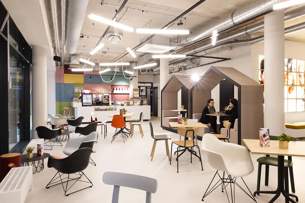 The bright communal area at the House of Hair featuring various seating options, including two small house-shaped structures used as private seating areas where two people are sitting. In the background, there is a counter with drinks and snacks.